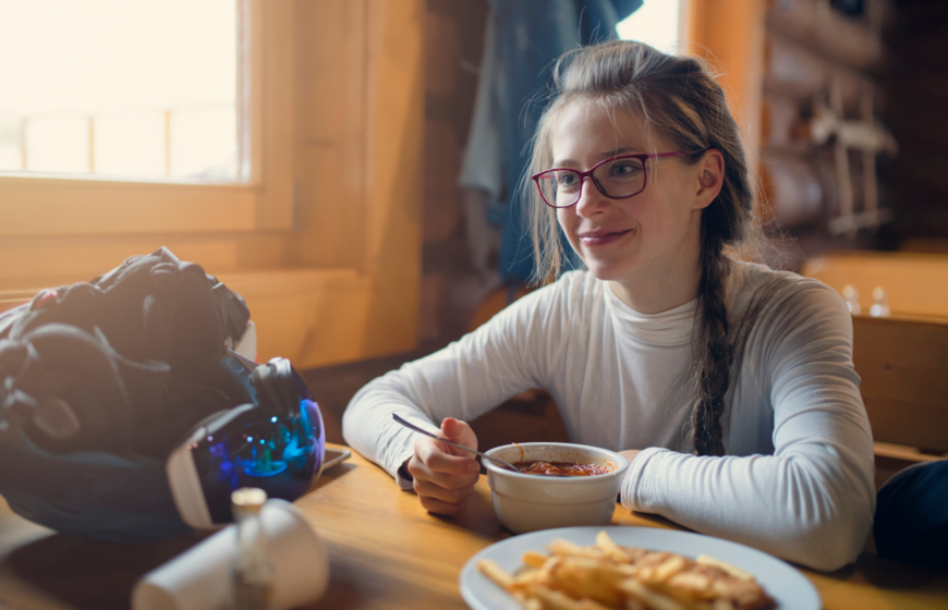 Young girl eating lunch at mountain restaurant with helmet and goggles on table