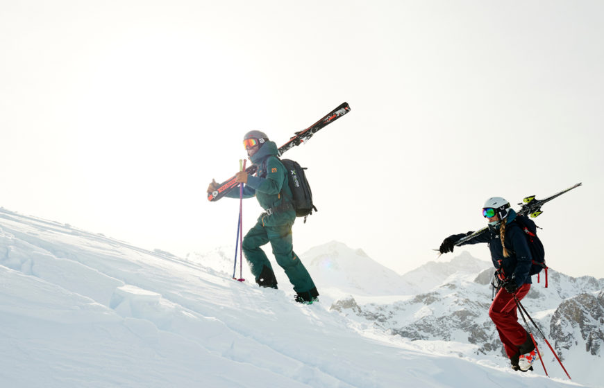 TDC ski instructor hiking up ski slope with female skier with skis on shoulders in Val d'Isere