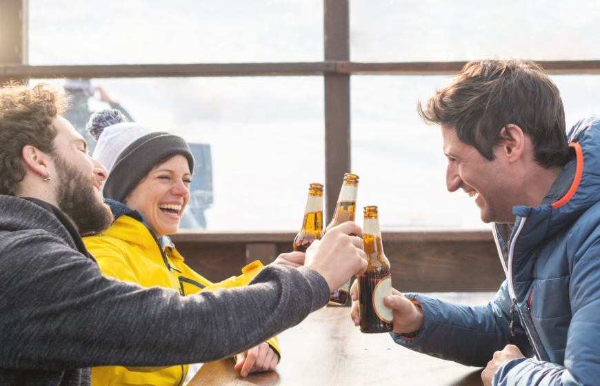 3 smiling friends chinking beer bottles on terrace of outdoor ski bar