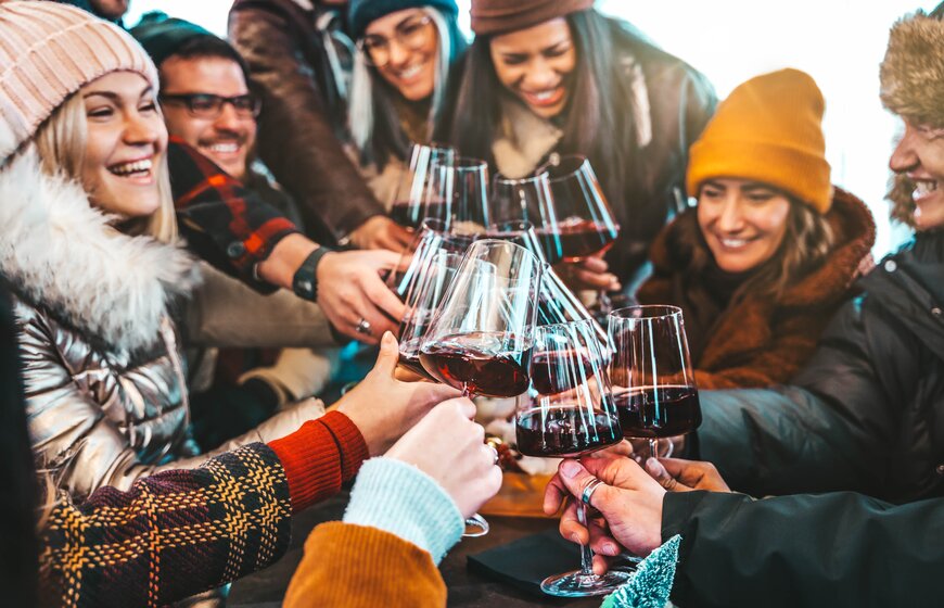 Group of friends dressed in warm jackets and hats saying cheers with glasses of red wine in outdoor bar