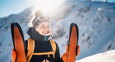 Smiling woman standing at top of snowy mountain holding up ski touring skis