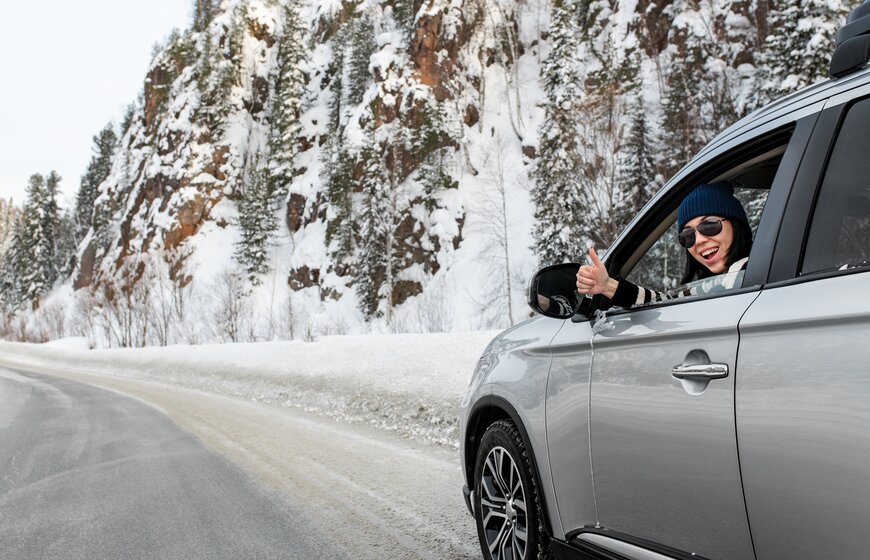 Smiling woman in silver car giving thumbs up through the window on a winding snowy mountain road