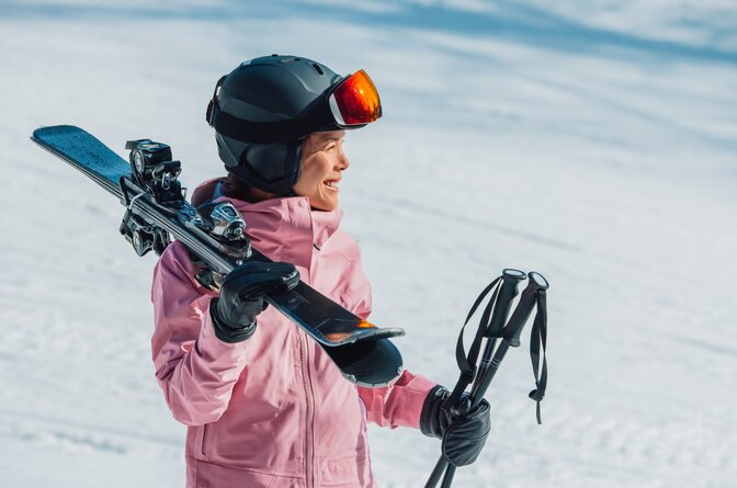 Smiling female skier in pink ski jacket and grey helmet carrying skis and poles