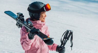 Smiling female skier in pink ski jacket and grey helmet carrying skis and poles