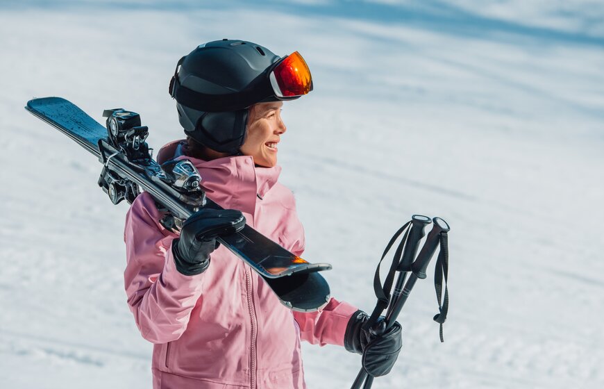 Smiling female skier in pink ski jacket and grey helmet carrying skis and poles