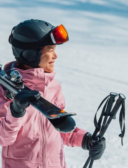 Smiling female skier in pink ski jacket and grey helmet carrying skis and poles