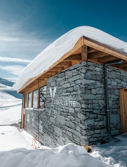 Exterior of L'Ouillette restaurant covered in snow on blue sky day in Val d'Isere