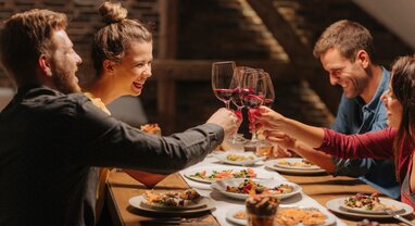 Two smiling couples clinking glasses of red wine together over table of food