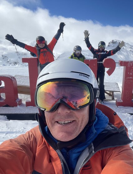 Henry Druce taking selfie in front of Val d'Isere letters with 5 skiers waving in background