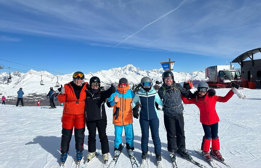 Oxygene ski instructor waving to camera with5 adult skiers at top of ski slope