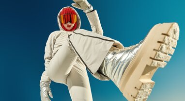 Woman standing over camera wearing beige ski outfit, gold snow boots and a red balaclava