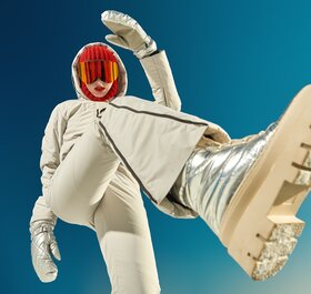 Woman standing over camera wearing beige ski outfit, gold snow boots and a red balaclava